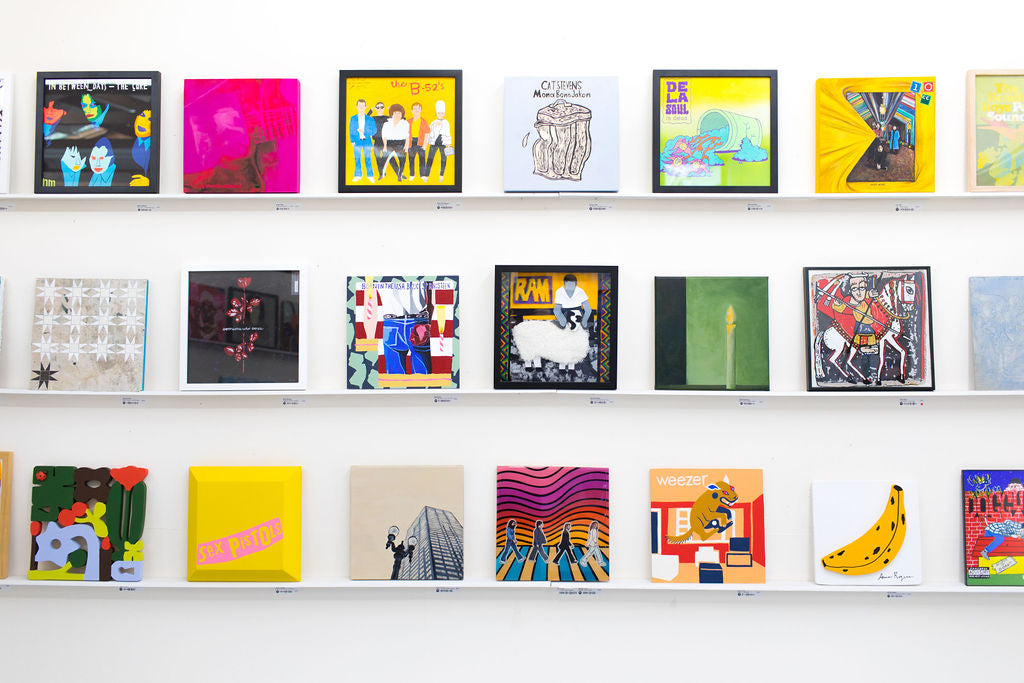 A white wall at WAX 2024 with about twenty reinterpreted album covers arranged on three picture ledges